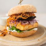 A burger with lettuce, square tofu, red onion, mushroom and fried carrot inside on a wooden round board and a small glass pot of white sauce at back. pot of