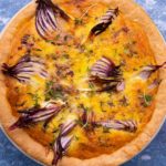 Top view of a round quiche with pieces of red onion peeping out on blue background.