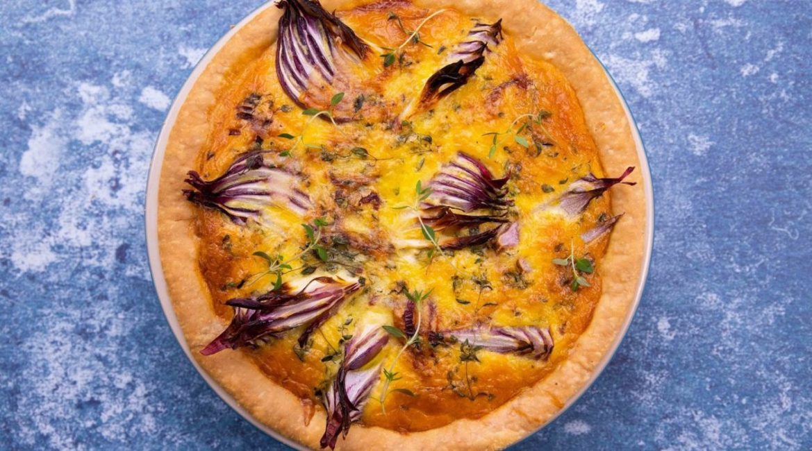 Top view of a round quiche with pieces of red onion peeping out on blue background.