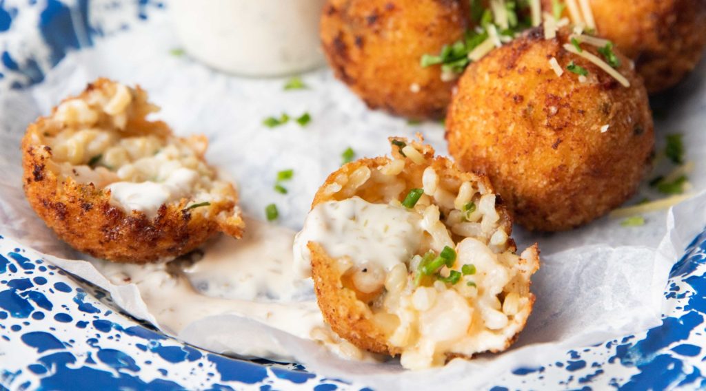 4 brown deep fried balls topped with herbs and one broke in 2 halves and a glass jar of white sauce on blue rimmed plate on blue grey background.
