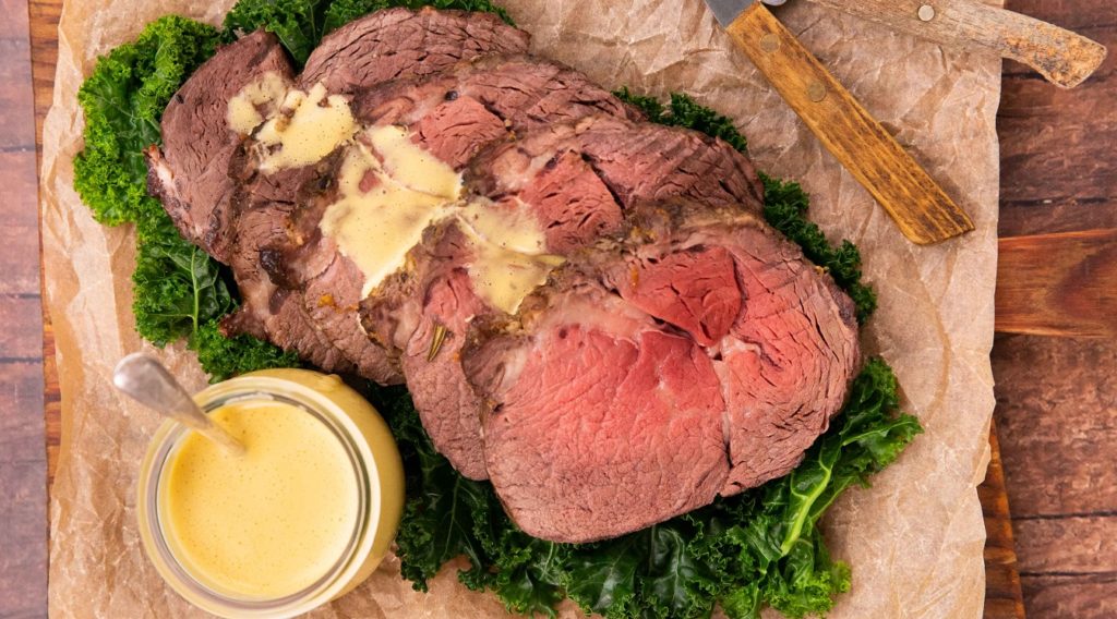 Roast beef slices on green on wooden board with yellow sauce and sauce pot.