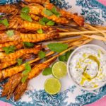 Cooked large prawn skewers on blue and white oval platter with lime halves and a pot of white sauce on pink background