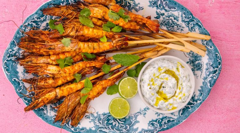 Cooked large prawn skewers on blue and white oval platter with lime halves and a pot of white sauce on pink background