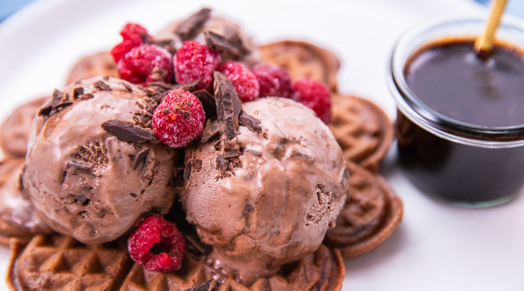 Chocolate waffles topped with ice cream balls, raspberries and chocolate shavings with a glass pot of chocolate sauce on white plate