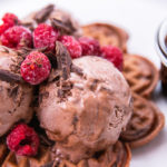 Chocolate waffles topped with ice cream balls, raspberries and chocolate shavings with a glass pot of chocolate sauce on white plate
