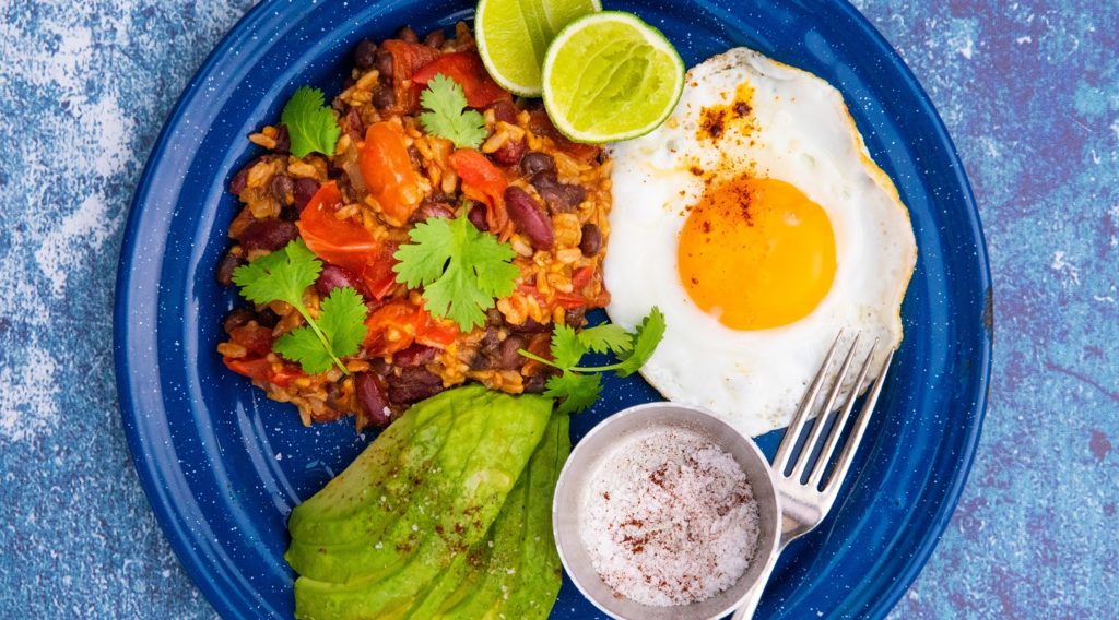 A fried egg, orange rice, avocado slices, lime halves a pot of red powder and a fork on blue round plate on light blue background