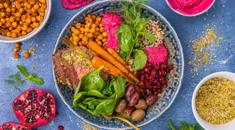 A large bowl of rice topped with colourful food, carrots, greens, pink paste, olives and red seeds and small dishes of ingredients around it.