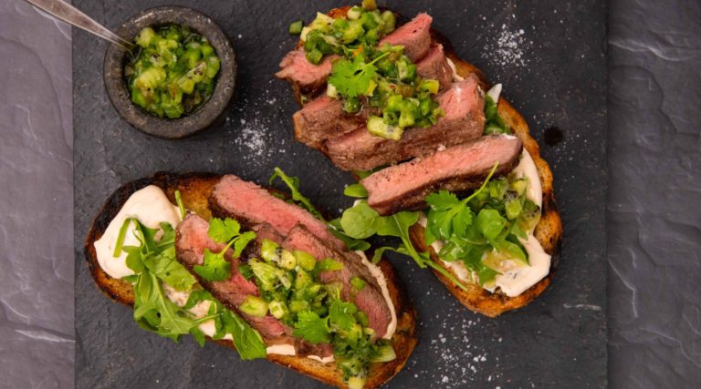 Two pieces of bread topped with cut steak, green salsa and white sauce under them on a stone slate with a small pot of green salsa.