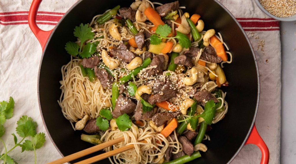 Beef, vegetables, cashews and noodles in a large two-handle orange dish with a pair of chopstick on white cloth.