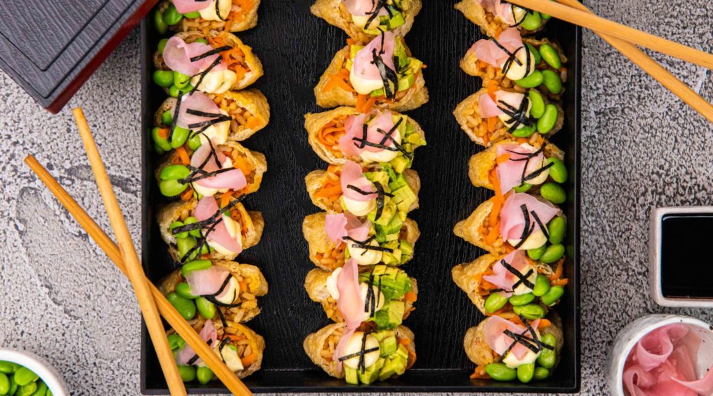 3 rows of 7 stuffed tofu pockets topped with pink and green food in black square box and two pairs of chopsticks, 3 small pots of pink, black and green food