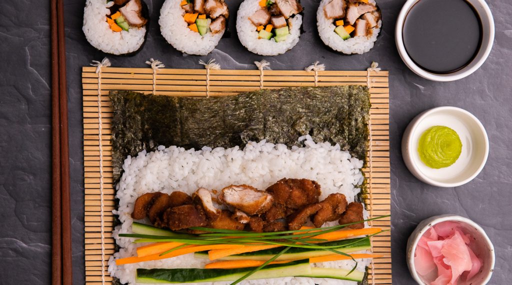 A nori sheet topped with rice and colourful chicken and vege fillings unrolled on bamboo mat. Four pieces of cut sushi on top, and three dishes of soy, wasabi and ginger on right.