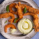 Deep fried crumbed prawns on a metal tray with a bot of pale green dipping sauce on blue background.