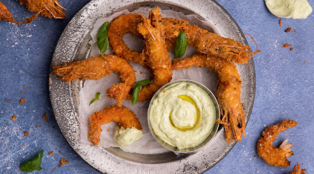 Deep fried crumbed prawns on a metal tray with a bot of pale green dipping sauce on blue background.