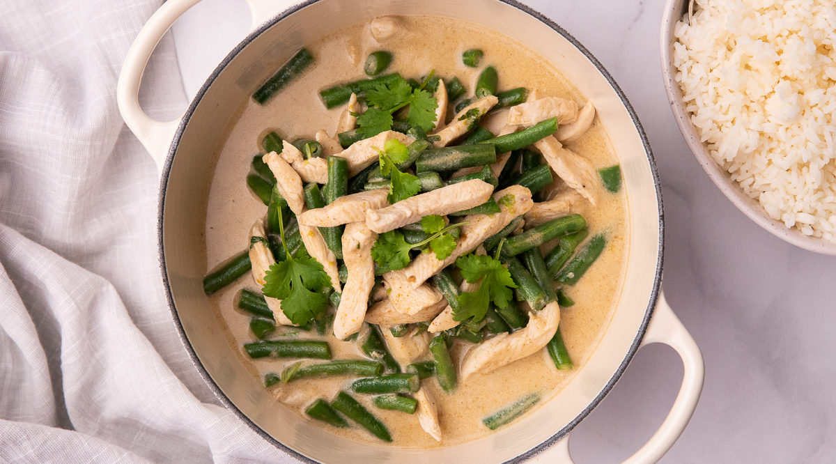 Creamy coloured soup with chicken pieces, green beans and herbs in a white double handled pan and a part view of white rice in a bowl on side.