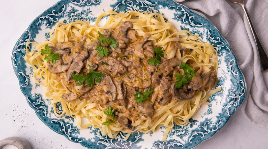 A platter of pasta topped with creamy mushroom sauce totted with green herbs on blue decorative platter.
