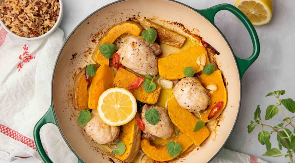 A green double handled casserole filled with chicken pieces, pumpkin and lemon half, and a bowl of reddish rice and oregano on white cloth