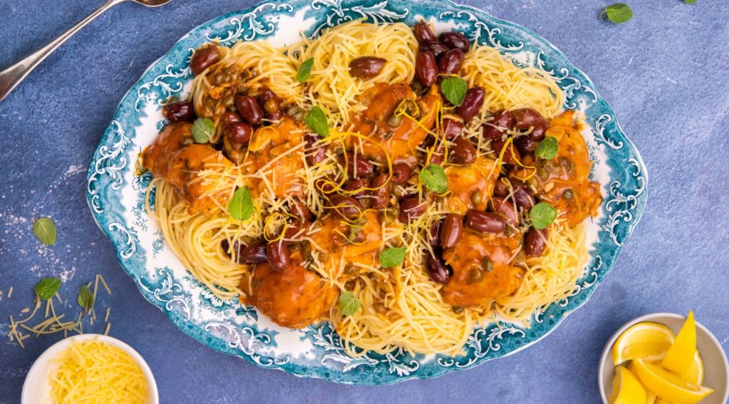 Spaghetti and tomato sauce covered chicken on a blue decorative platter with black olives and herb scattered on top on blue background and small dishes of cheese, lemon etc.