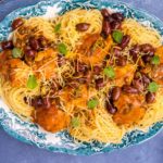 Spaghetti and tomato sauce covered chicken on a blue decorative platter with black olives and herb scattered on top on blue background and small dishes of cheese, lemon etc.