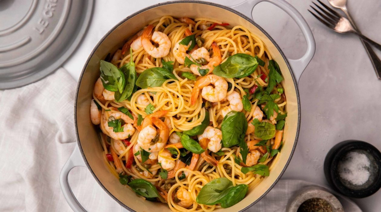 A grey double handled pan full of pink spaghetti with prawns and herbs. 2 forks, pan lid and 2 small bowls of salt and pepper on white background.
