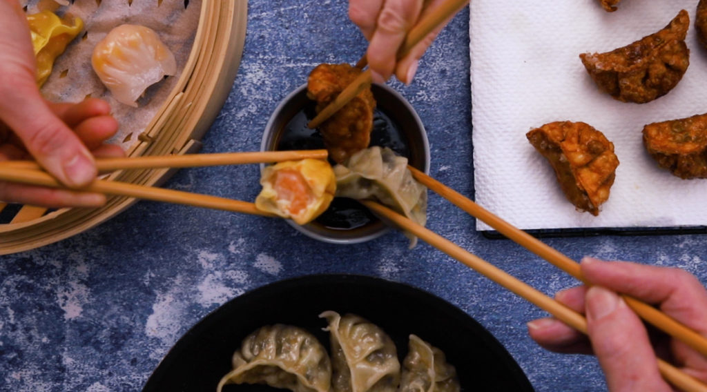 3 hands each dipping a dumplings cooked in 3 different ways using chopsticks, more dumplings shown around them.