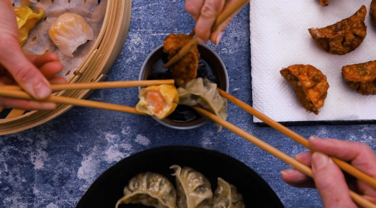 3 hands each dipping a dumplings cooked in 3 different ways using chopsticks, more dumplings shown around them.