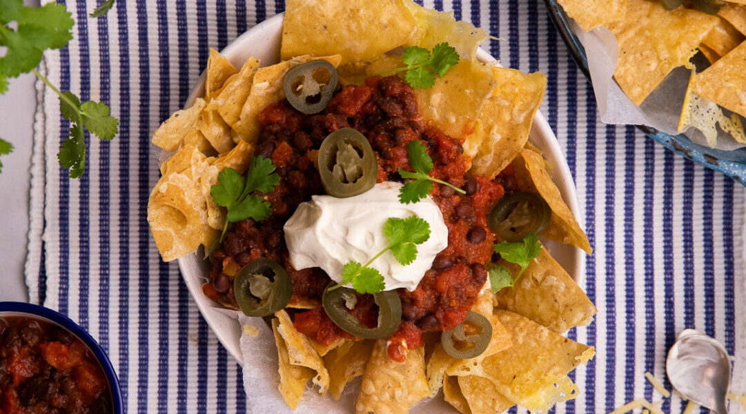 On a blue striped mat, a bowl of corn chips topped with red black chilli, a blob of white cream, pieces of green chilli and herb. Ingredients in corners outside of the bowl.