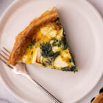 A slice of green and white quiche on a white round plate with a fork. A small part of the whole quiche shown on one corner with a pink cloth.