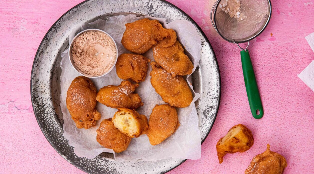 10 golden brown fried donut and a small pot of sugar on a metal plate on pink background with 2 more donut and a tea strainer at side.