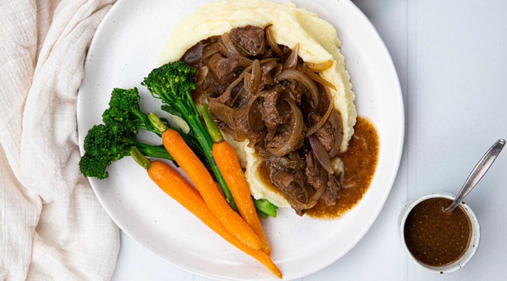 A plate of dark brown beef stew on potato mash with baby carrots and green vegetable on a white plate and a small pot of dark sauce.