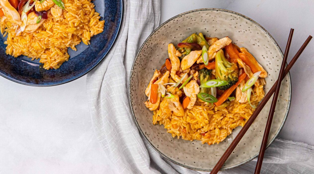 2 bowls of orange coloured rice with stir fried veges and chicken at side, one bowl with chopsticks on fabric.