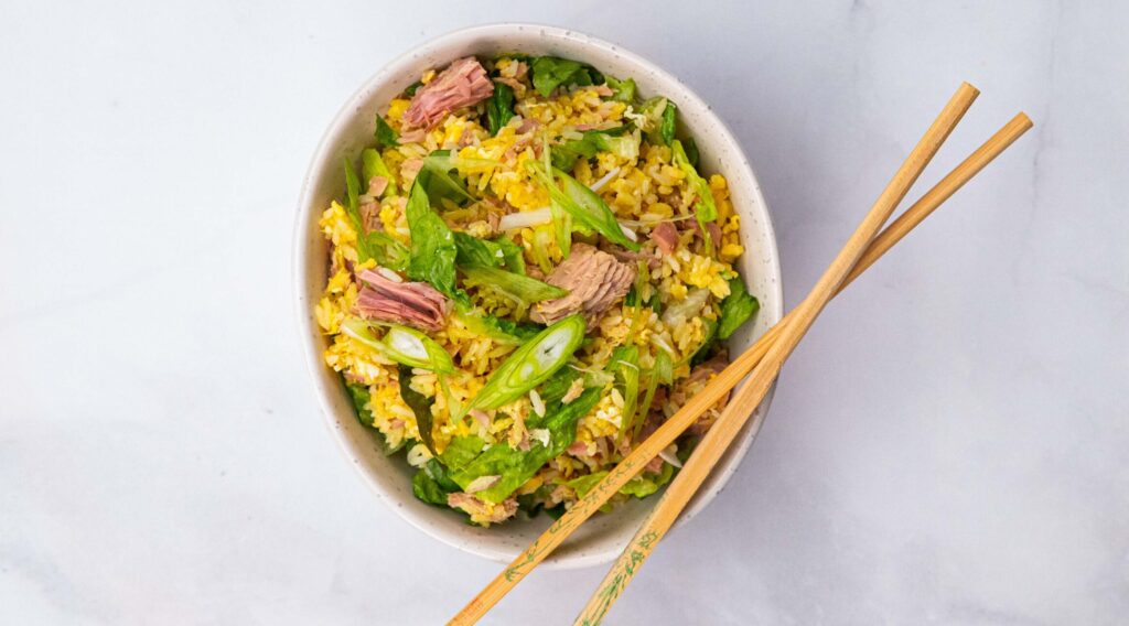 A bowl of yellow fried rice mixed with pink tuna meat, lettuce and spring onion slices and a pair of wooden chop sticks