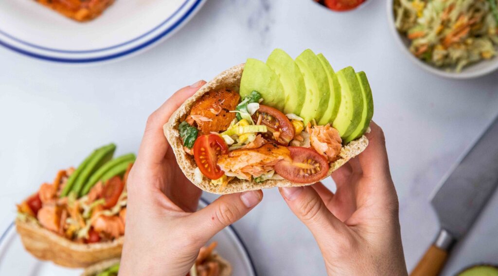 Hands folding a pita bread filled with tomato, salmon and avocado.