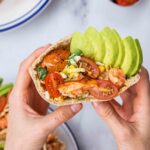 Hands folding a pita bread filled with tomato, salmon and avocado.