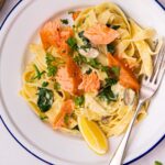 A pasta dish with salmon, greens and lemon on a blue rimmed plate with a fork on white board and cheesy toasts at sides.