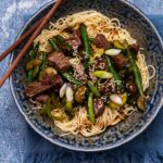 Noodle topped with beef and greens stir fry in a bowl with chopsticks on blue surface.