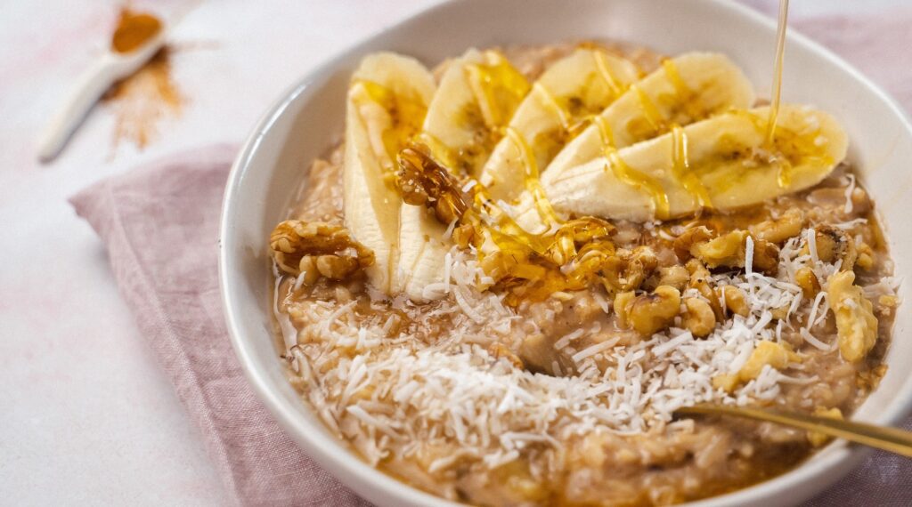 A rice pudding in a white bowl topped with banana slices, walnuts a,d coconut shavings, drizzle of honey