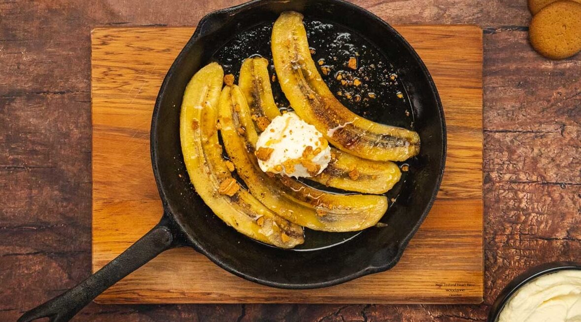4 long slices of bananas cooked in a black skillet topped with white cream drop, honey drizzle and crumbs