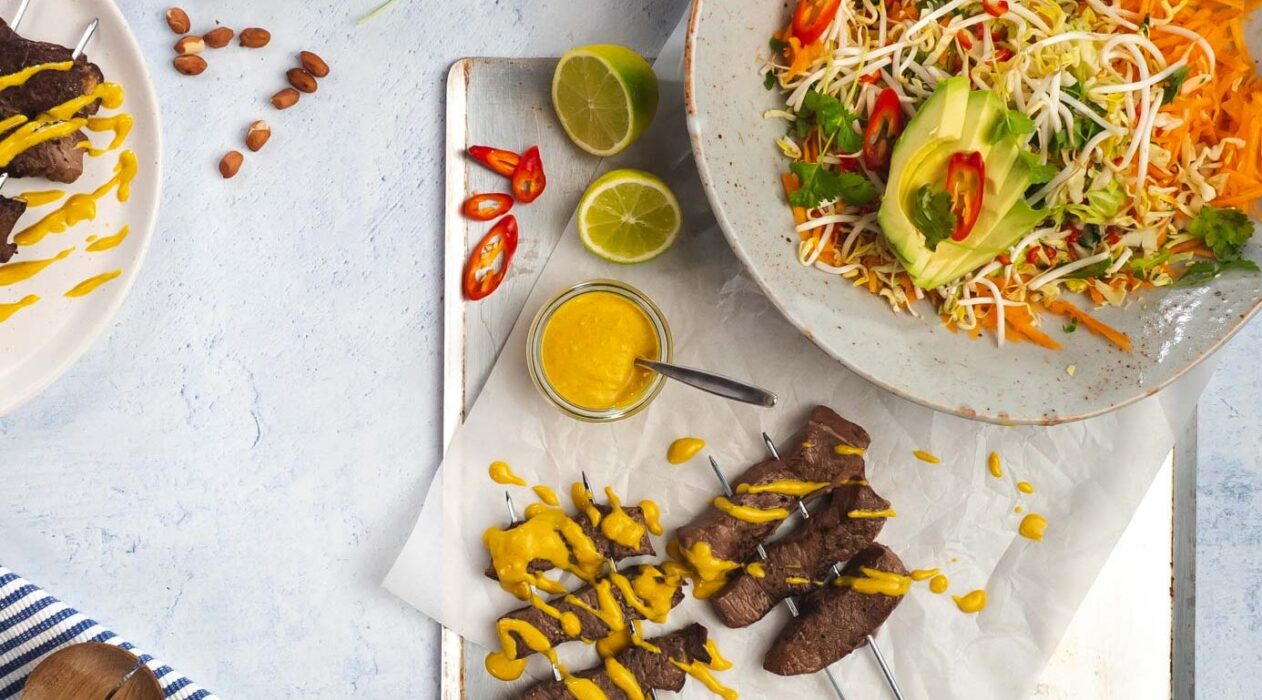 Skewered beef drizzled with yellow sauce on a tray with a bowl of colourful salad, lime halves and chilli.