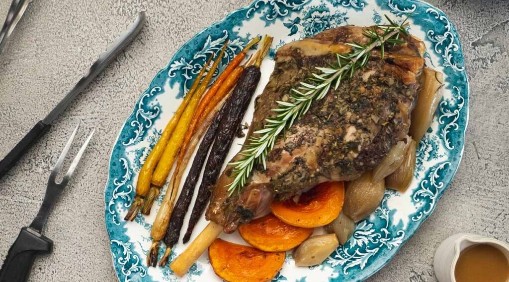 Roast leg of lamb on blue decorative plate with roast vegetables and fork tongs