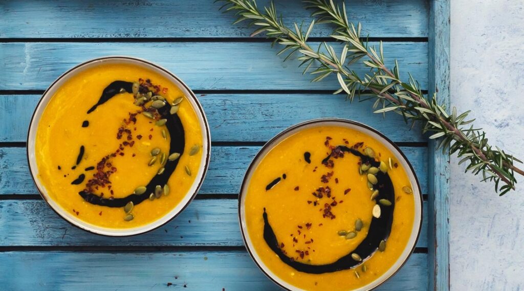 2 bowles of orange coloured soup topped with black swirl, green seeds and red flakes on blue tray with sprigs of rosemary