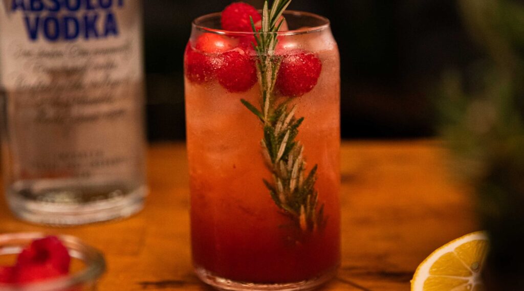 A tall glass of pink drink with rosemary and raspberries on wooden board, lemon half and a bottle of vodka