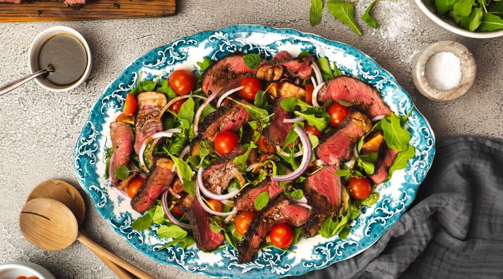 Sliced beef, green leaves, tomatoes and onion salad on oval blue platter surrounded by salad server, pots of salt Pepper