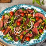 Sliced beef, green leaves, tomatoes and onion salad on oval blue platter surrounded by salad server, pots of salt Pepper