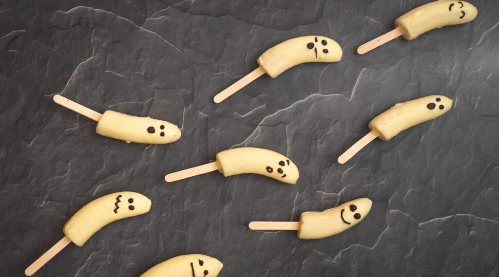 8 white chocolate coated banana halves with faces on popsicle sticks on dark stone board.