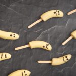8 white chocolate coated banana halves with faces on popsicle sticks on dark stone board.