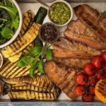 Grilled meat, courgette and aubergine slices, tomatos and orange on a metal tray with a bowl of green salad