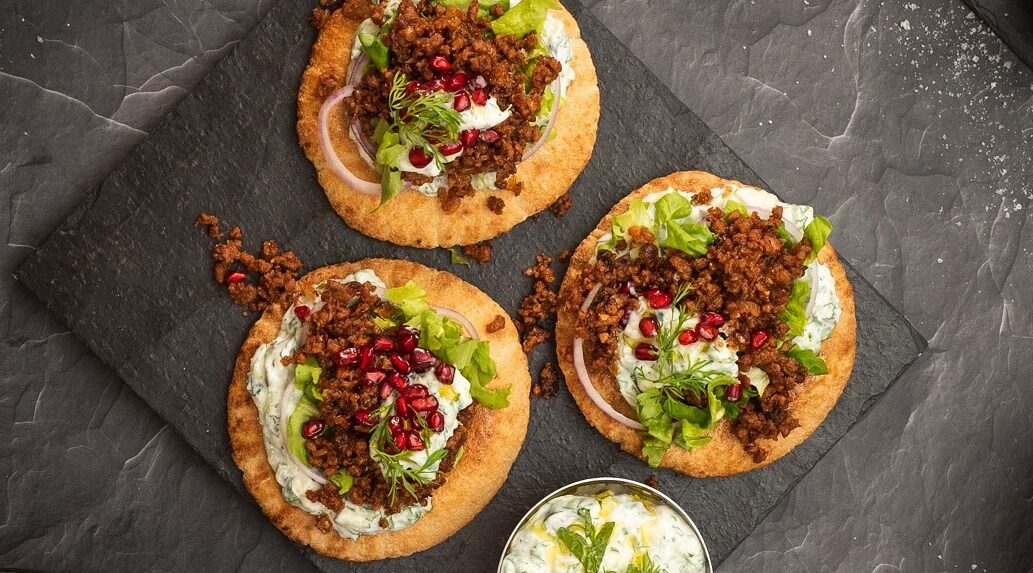 3 round bread topped with white sauce, mince, lettuce and red berries on stone board..