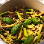 Penne pasta, mince, basil leaves and asparagus in a pot on white board.