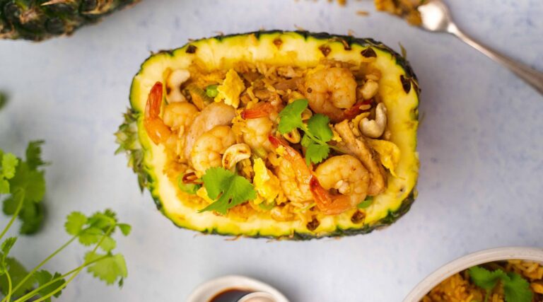 Fried rice with prawns, eggs and coriander in a pineapple half shell on white board with a fork and dishes.