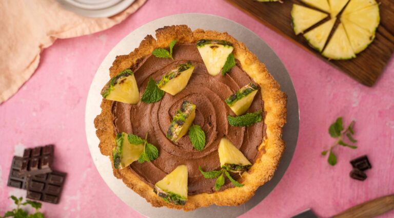 op view of a round chocolate tart with pineapple pieces on pink background with cut pineapple on wooden board, chocolate pieces and plates.
